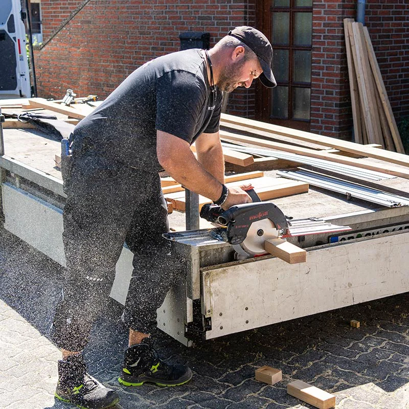 Man in a black cap and T-shirt uses a power saw to cut a wooden beam on a workshop trailer outdoors, wood shavings flying around.