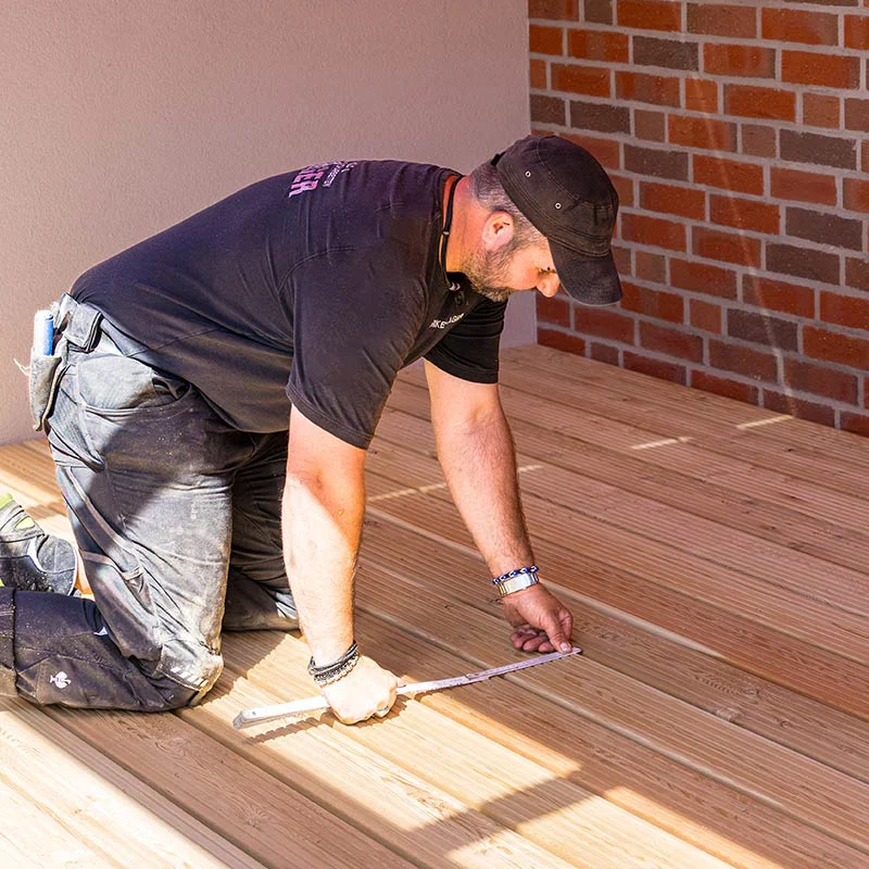 Carpenter kneels on a wooden deck, measuring with a tape measure against a brick wall backdrop